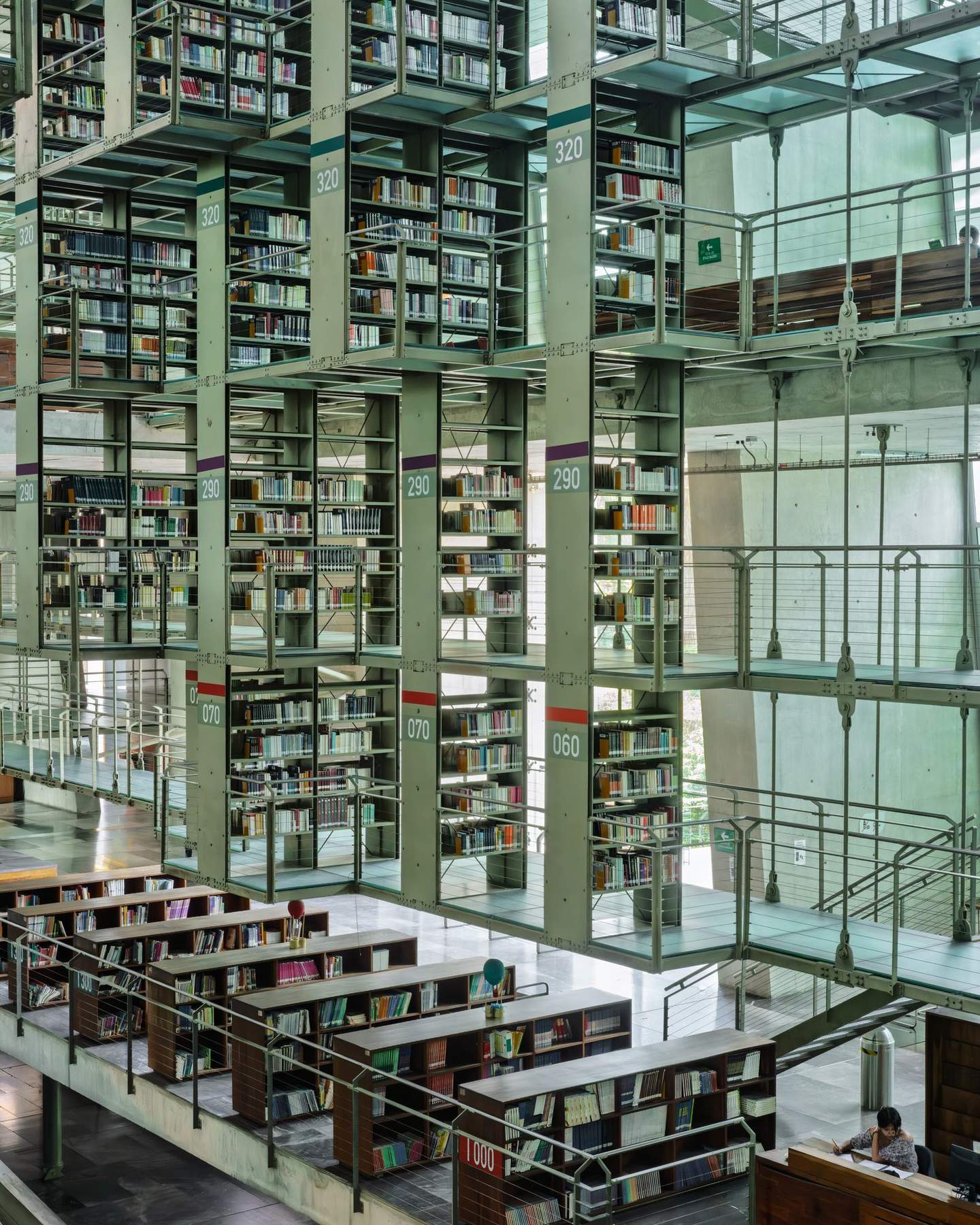 Biblioteca Vasconcelos central nave from upper walkway, staircase receding to warm wooden reading room terminus, two figures at different levels