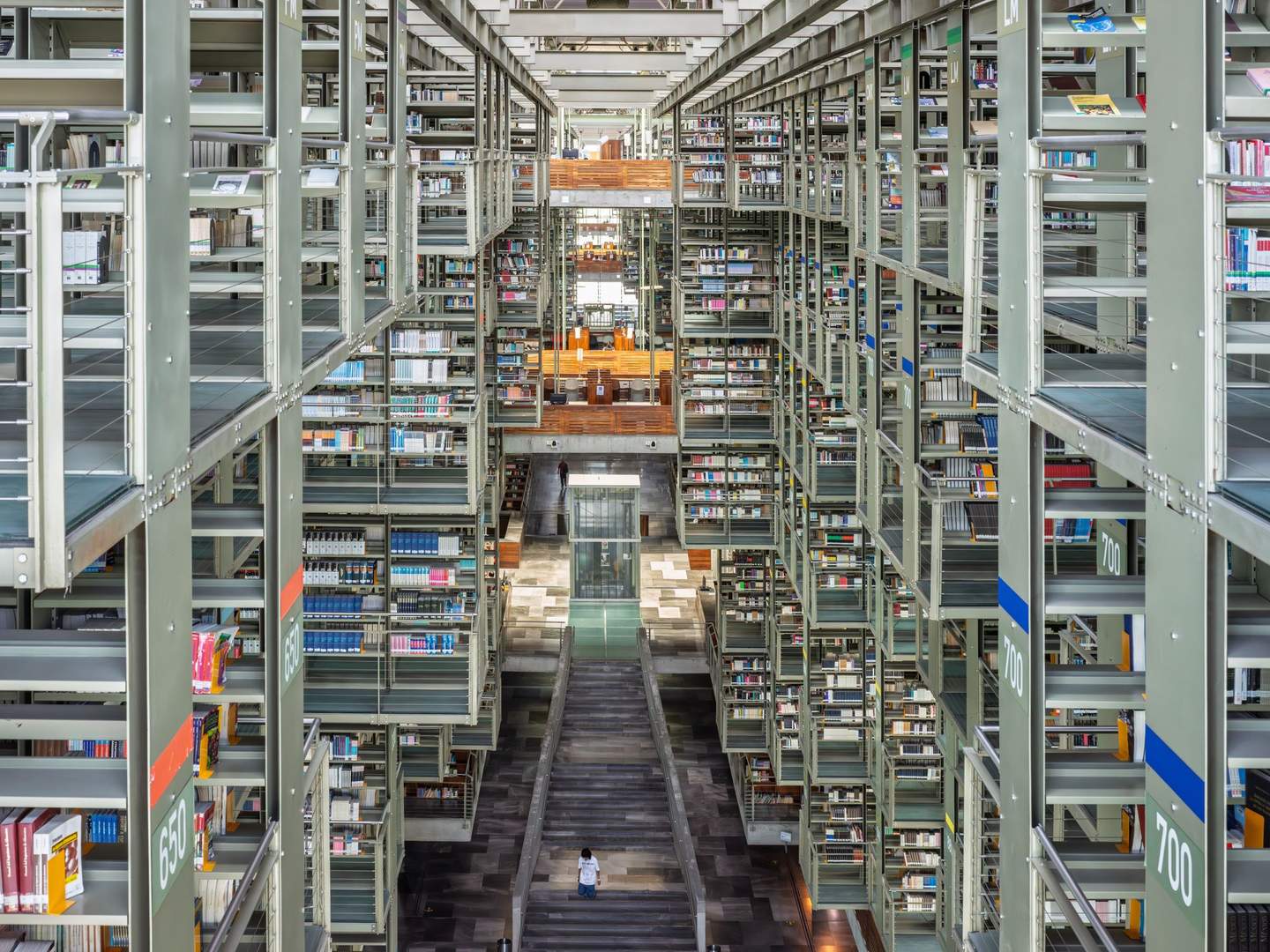 Looking up through full height of Biblioteca Vasconcelos suspended stack system, figure in motion on mid-level walkway, stacks receding upward into roof structure