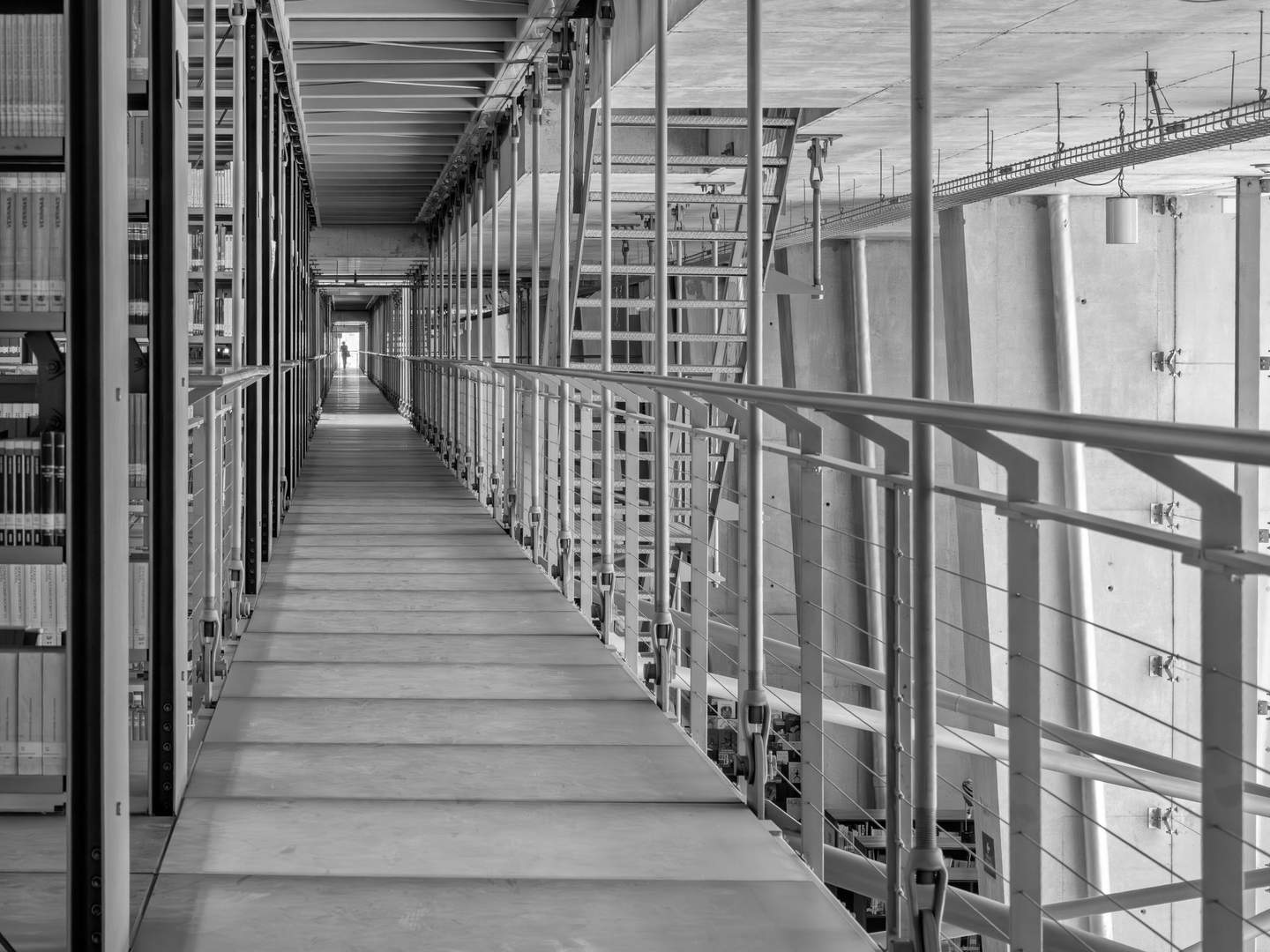 Biblioteca Vasconcelos suspended walkway corridor in black and white, figure at vanishing point, roof truss and suspension hardware above, book spines lining left edge
